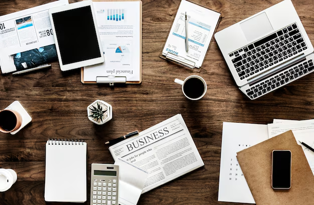 A top shot of a laptop, a tablet, a phone, cups of coffee, a calculator, notepads, and charts