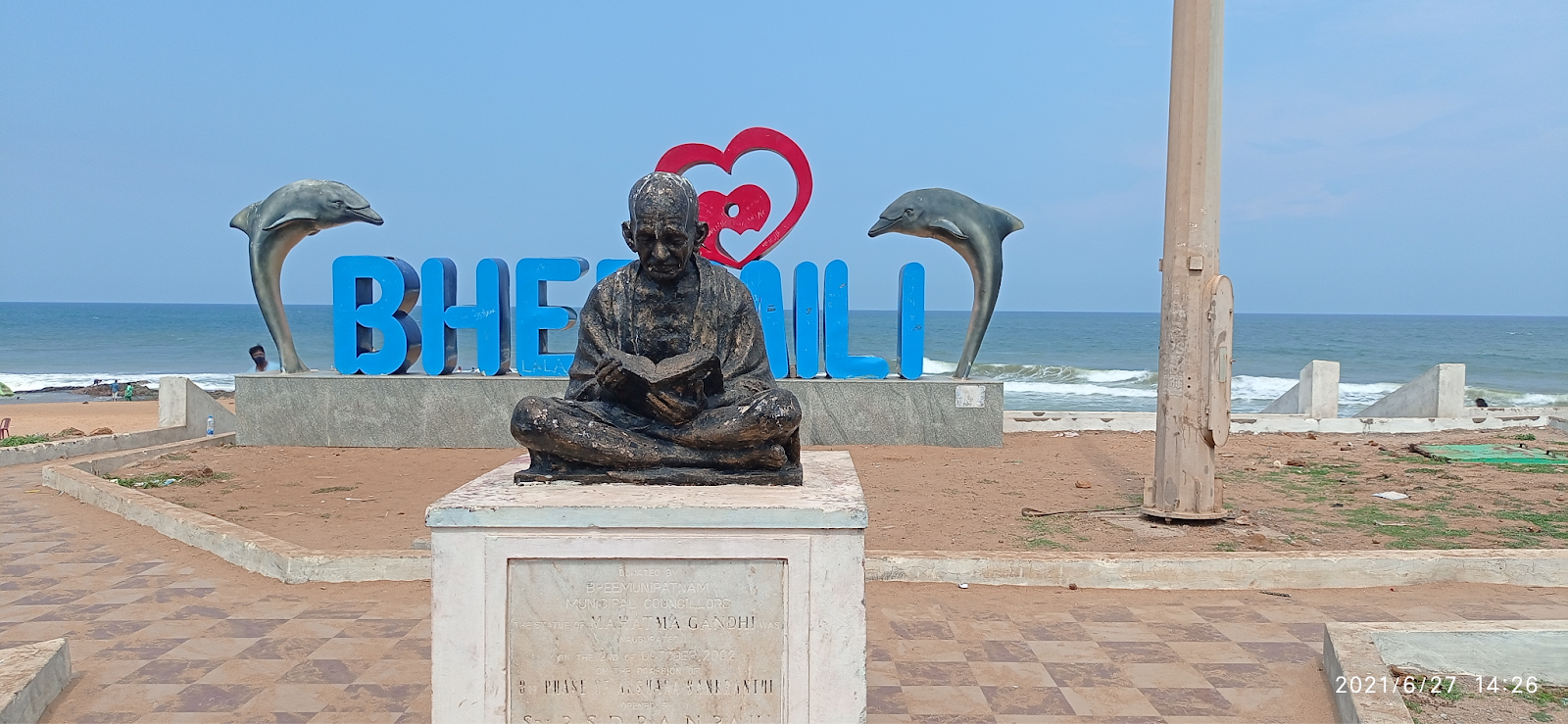 A statue of Mahatma Gandhi sitting on a pedestal on the shore of Bheemili beach. Two dolphins are sculpted on the beach, and a heart is placed between them. The statue has waves crashing in the background.