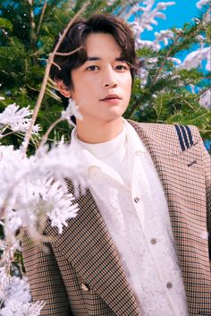 This contain Minho in a suit standing next to some white flowers and trees with blue sky behind him