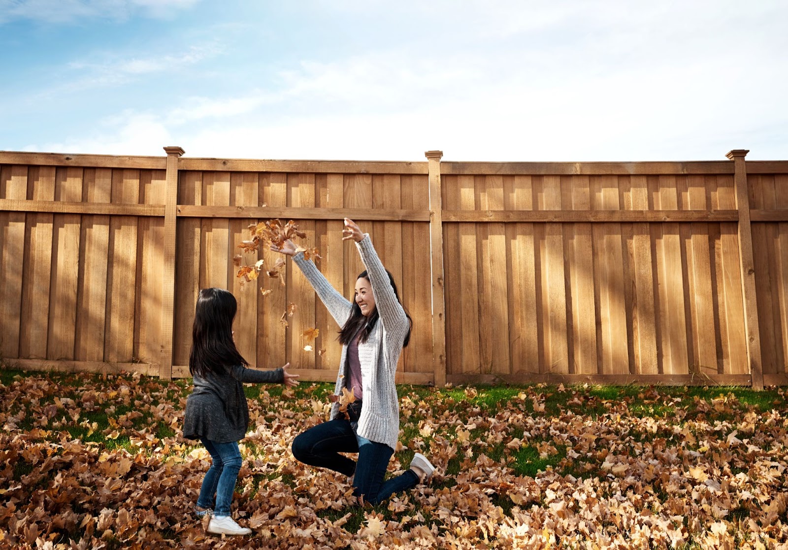 A wooden perimeter fence encircles a property where a mother and daughter joyfully play with dried leaves.