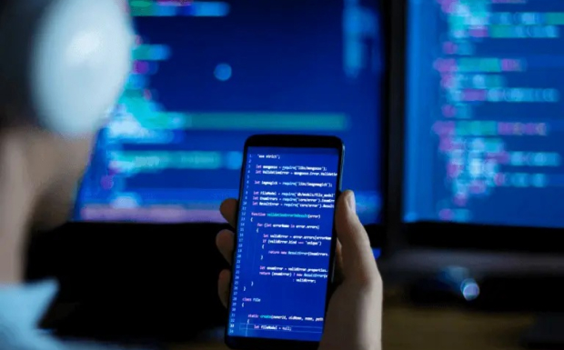 A developer working on a mobile app, with their smartphone showing the app's code and their computer monitors displaying additional development tools for Mobile App Development Process
