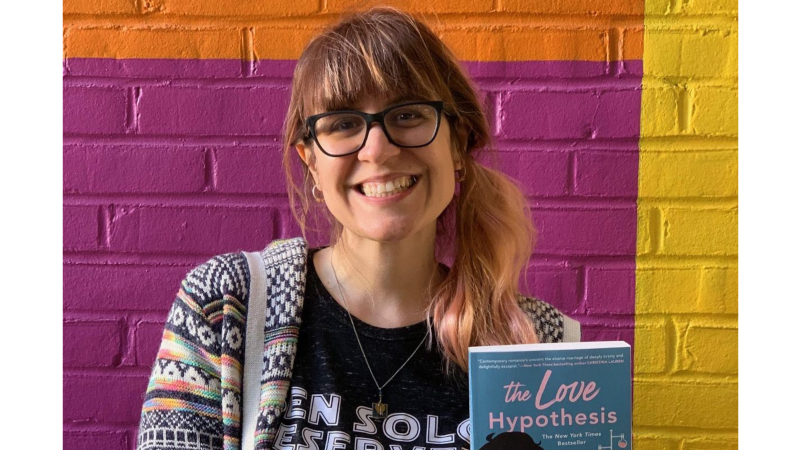 Author Ali Hazelwood smiling while holding the book The Love Hypothesis, standing against a colorful wall with orange, pink, and yellow.