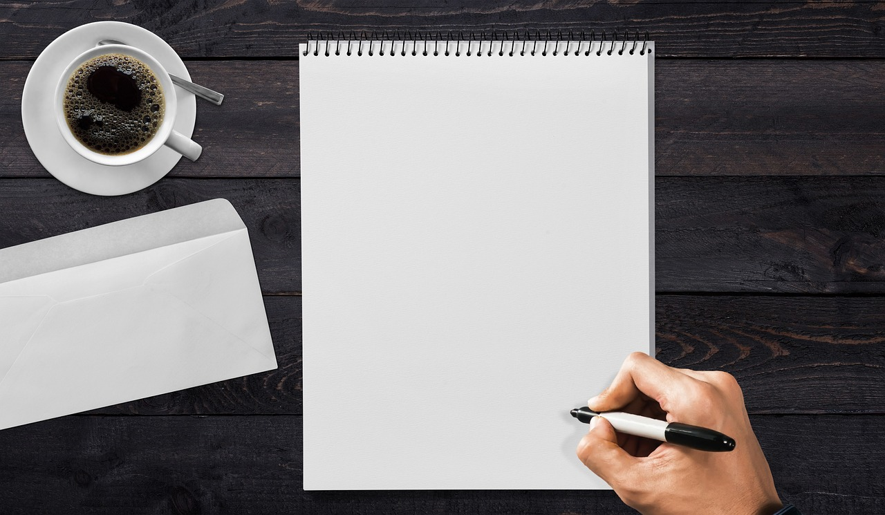 dark wooden office desk with coffee, notebook, envelope and man writing resignation on empty scheet of paper