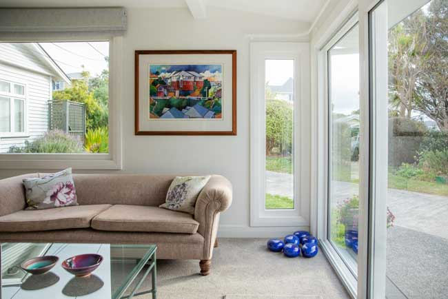 Bright living room with a beige sofa, glass coffee table, and a colorful painting on the wall. Large uPVC windows overlook a garden. Blue decorative items are on the floor.