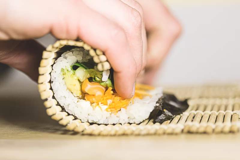 A close-up of a hand rolling sushi with a bamboo mat, showing fresh ingredients like rice, avocado, and vegetables inside the nori sheet.