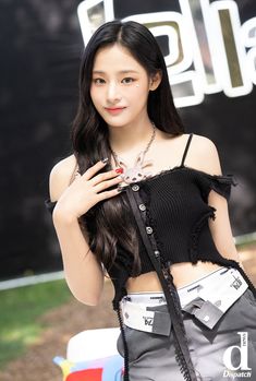 This contains NewJeans  Minji wearing a black crop top and an ash colour skirt with her hands on her chest