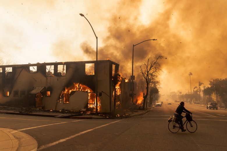 Flames and smoke can be seen coming out of a building as businesses in the Altadena area burn due to the Eaton Fire. Nearby, a person rides their bike as burnt debris float in the sky as smoke fills the atmosphere.