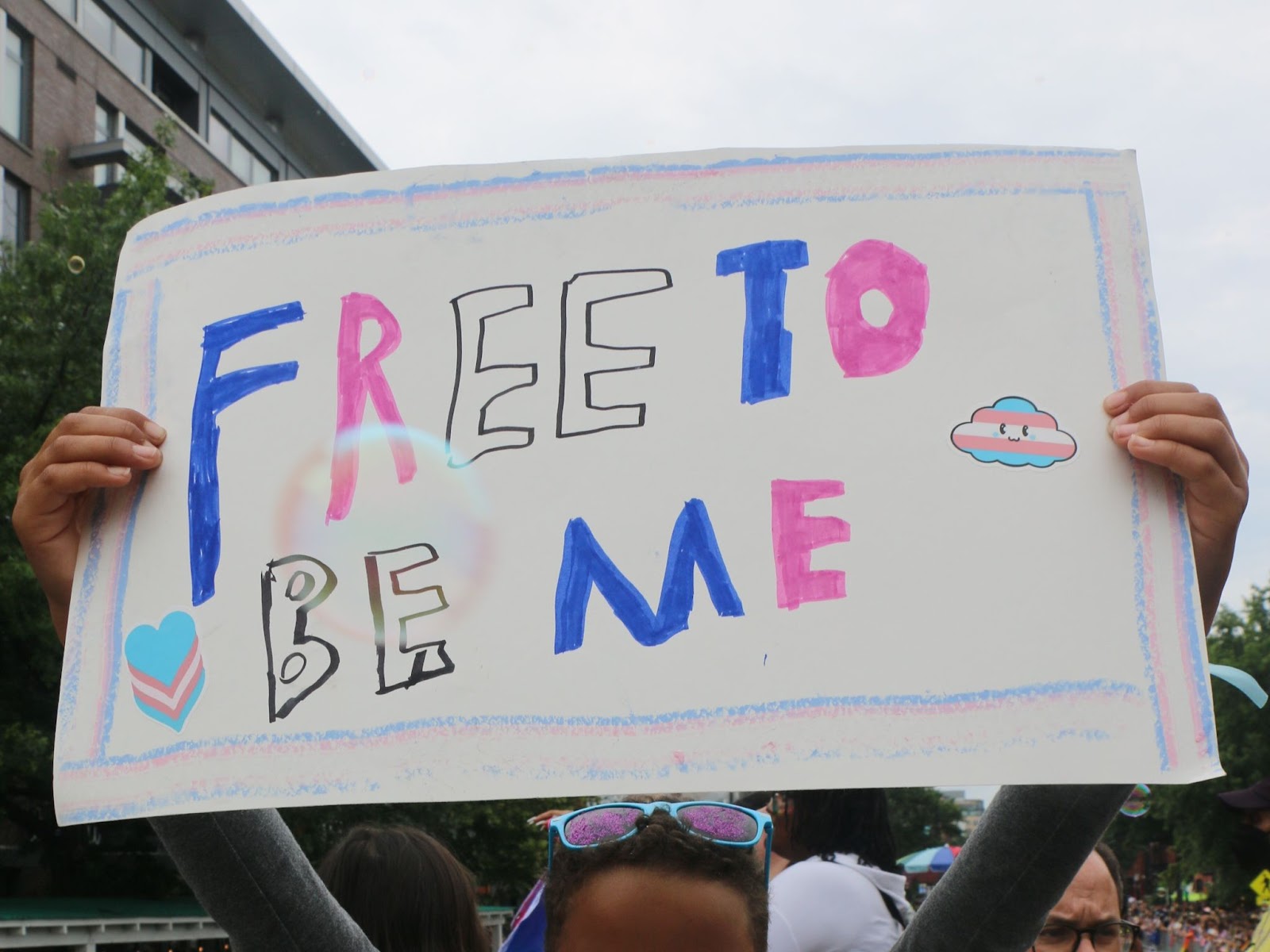 A person holds a blue, pink, and white poster that says Free To Be Me.