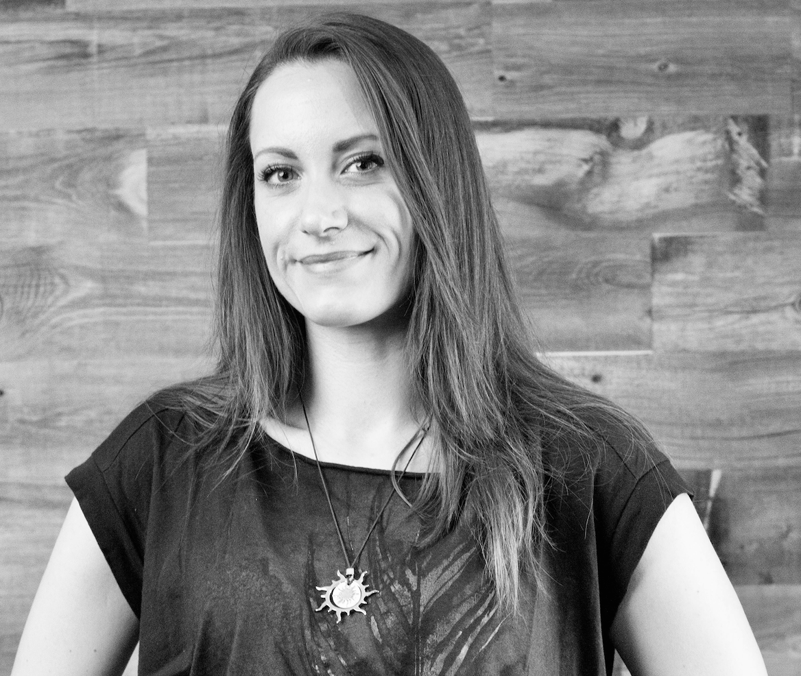  a black and white headshot of Antonella as she stands in front of a wooden wall, facing the camera and hinting a smile. Antonella is a white woman with long dark hair. She wears a dark t-shirt with a feather printed on it, and a necklace of a tribal sun. 