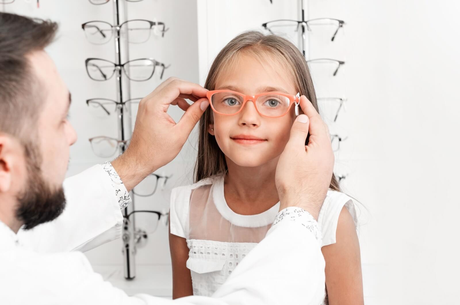 An optician placing a pair of eyeglasses with an orange frame on a smiling girl's face.