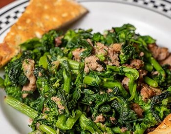 A plate of sautéed broccoli rabe with crumbled sausage, served with crispy flatbread on a white dish with a black checkered rim.