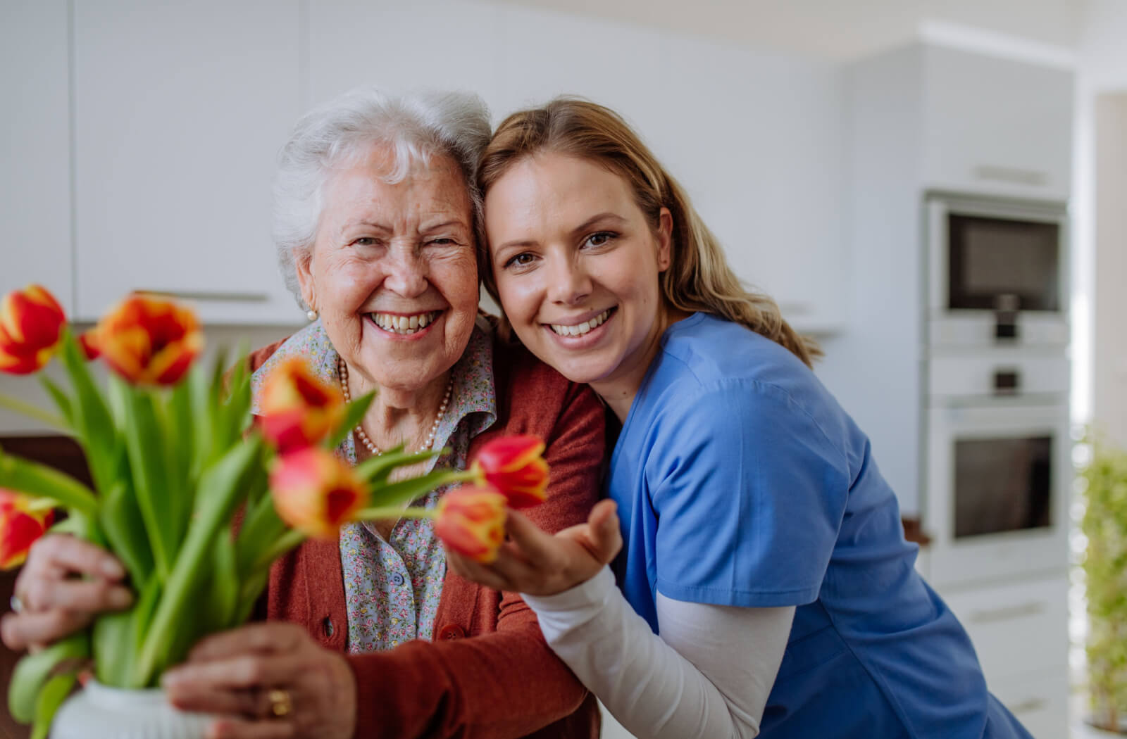 A caregiver hugging an older adult in front of a bouquet of tulips in assisted living.