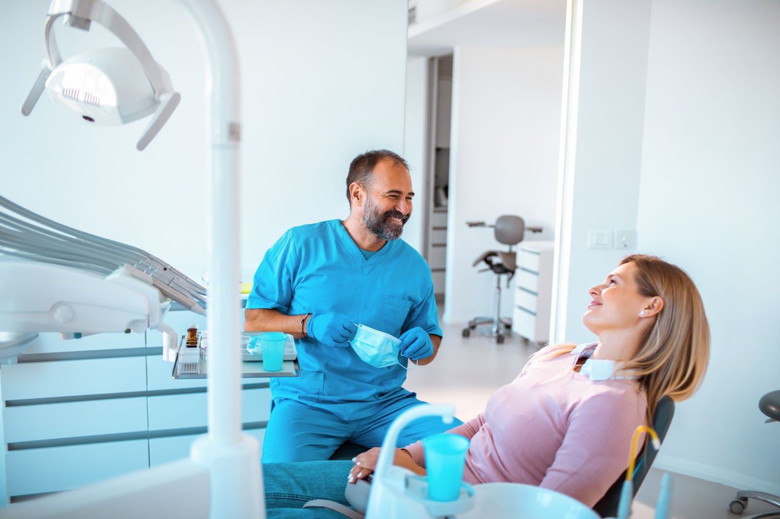 A woman sitting back in a dental chair and smiling at her dentist as he smiles back at her and holds a medical mask in his gloved hands, about to put it on.