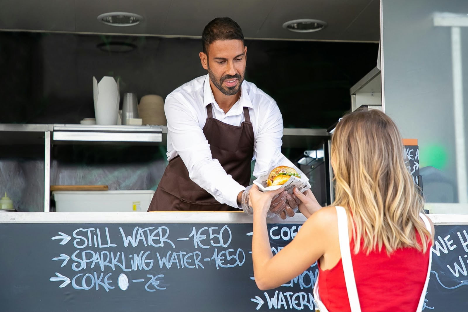 A food truck vendor hands a sandwich to a female customer at the service window.