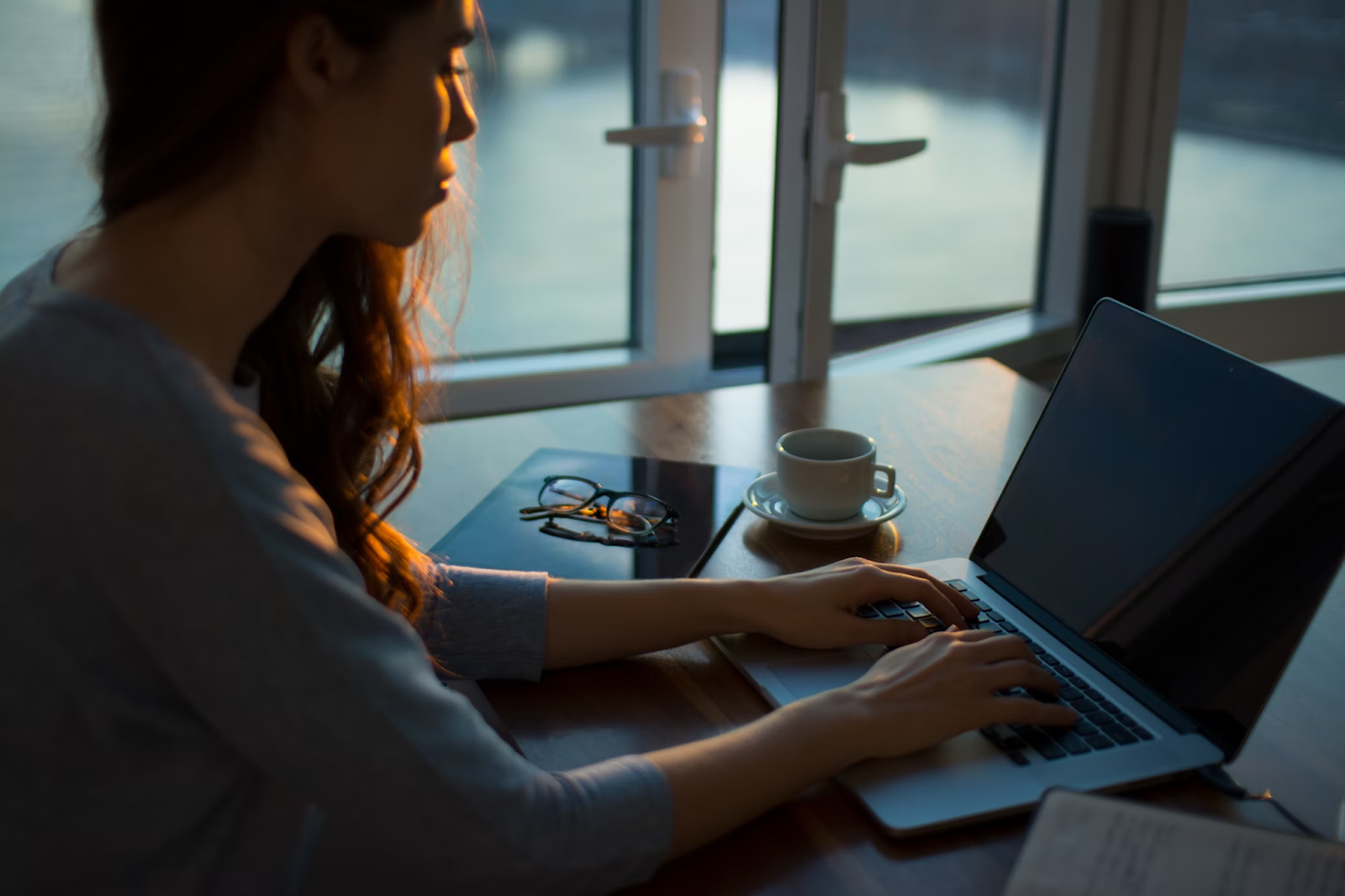 Close-up of a woman typing on a laptop about writing a professional short bio, with coffee and eyeglasses nearby, in an evening setting