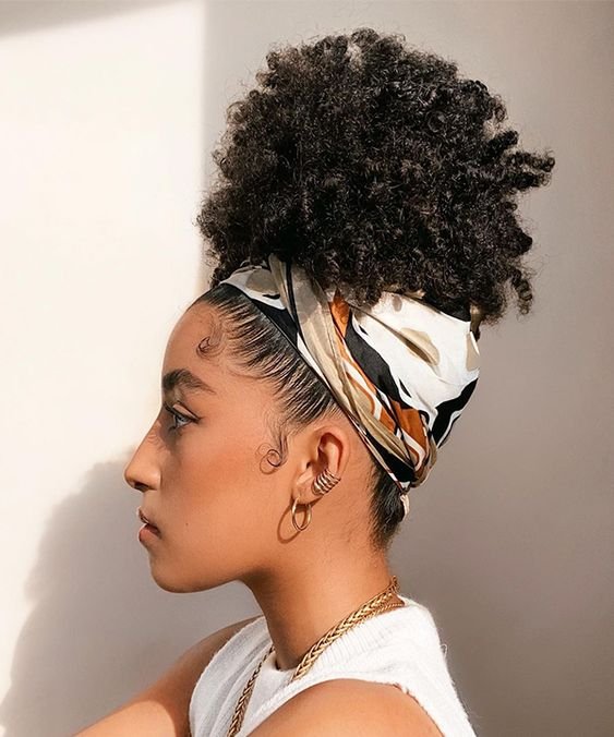 Mulher negra usando o cabelo crespo preso no alto da cabeça com um lenço branco com detalhes em cores terrosas. De perfil, ela usa uma blusa branca, um colar e brincos dourados.