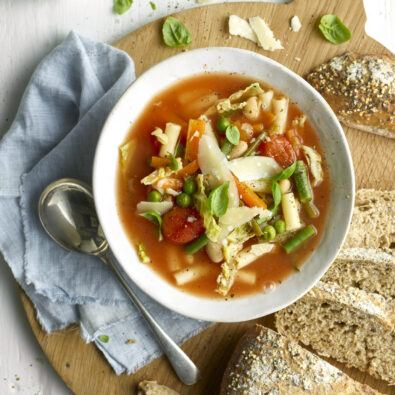 A bowl of homemade minestrone soup filled with vegetables, pasta, and herbs, served on a wooden board with slices of seeded bread and a silver spoon.