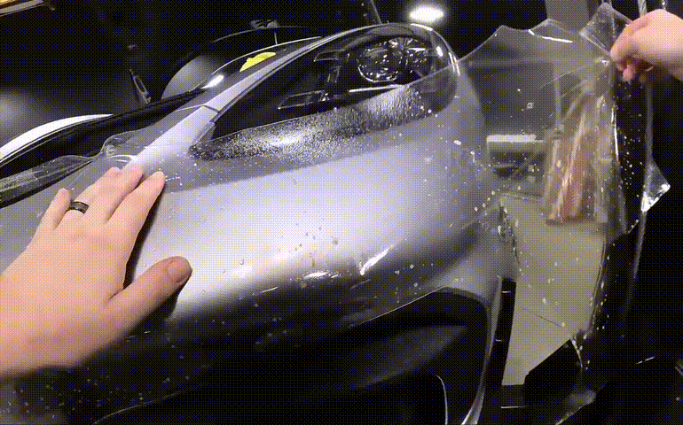 Close-up of a person carefully smoothing out a paint protection film on a sleek silver car's hood. The film application process includes precision and attention to detail, with water droplets visible for proper alignment