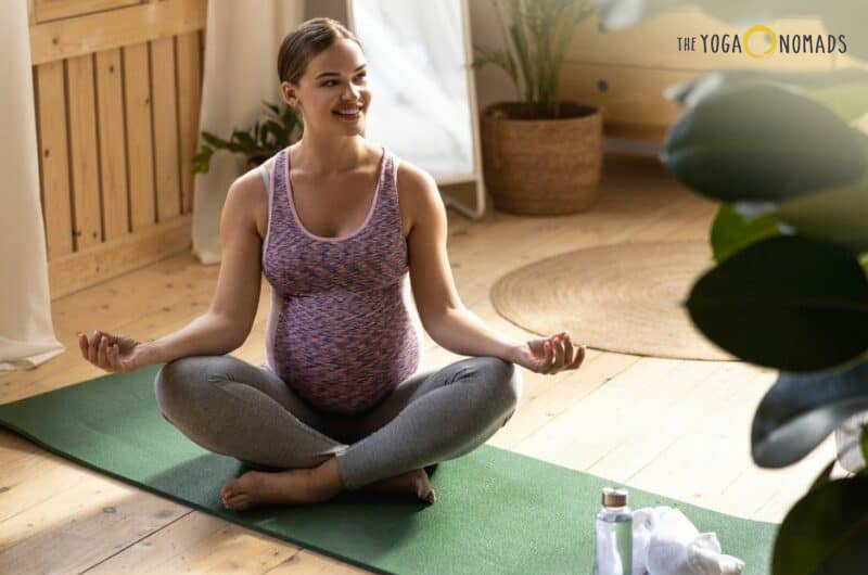 An individual sitting on a green yoga mat in a cross-legged position with hands resting on the knees in a meditative pose. The setting appears to be a bright, sunlit room with wooden flooring. There is a water bottle and some personal items to the side of the yoga mat.