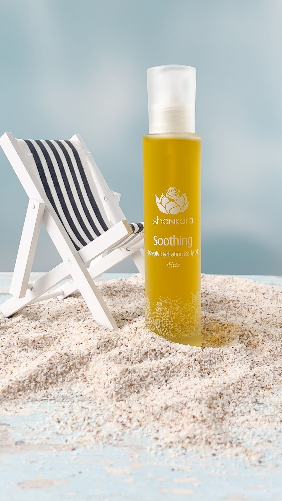 Close up image of Shankara Naturals Soothing Body Oil on sand next to a miniature striped black and white sunlounger chair. Featured in the blog Luscious Locks: How Body Oil Can Transform Your Hair Care Routine