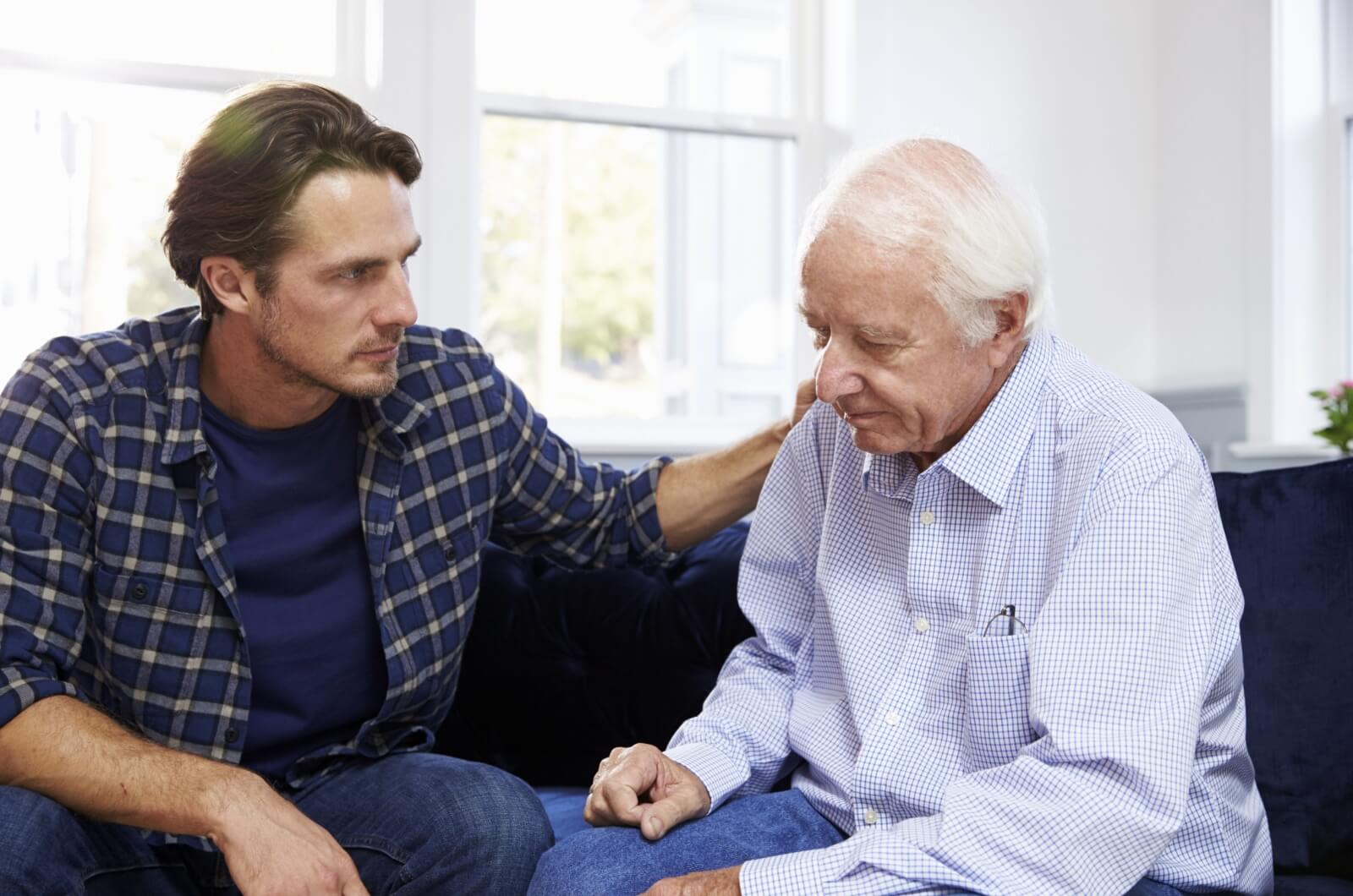 An adult child reassuring a parent with dementia by placing a hand on their shoulder during a conversation.