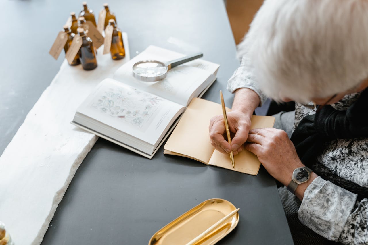 An elderly person writing in a notebook with a gold pen, surrounded by an open book, a magnifying glass, and small bottles, suggesting a research or study session.