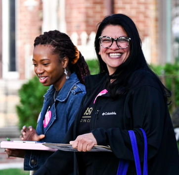 Rashida smiles while holding a clipboard while walking with a young Black woman