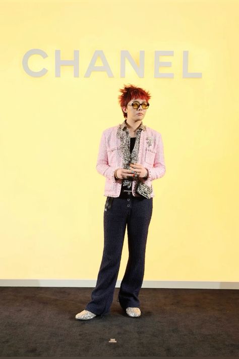 This contains an image of G-Dragon with red hair and glasses standing in front of a chanel logo on a yellow wall