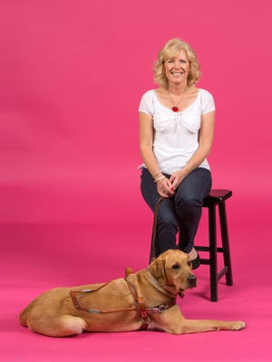 The image shows a woman with blonde, shoulder-length hair sitting on a black stool in front of a bright pink backdrop. She is smiling, wearing a white top and dark pants. In front of her, lying on the floor, is a guide dog with a golden-brown coat, wearing a harness, indicating that it is a working dog.