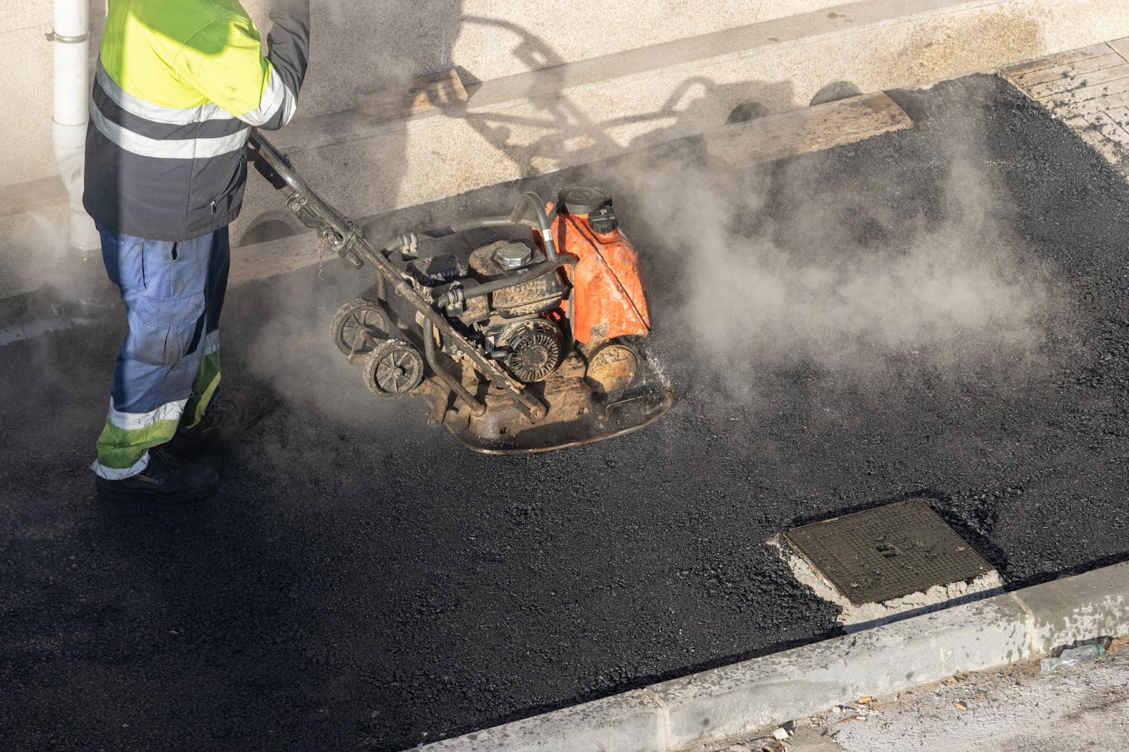 A worker operates a vibrating plate compactor machine smoothing freshly laid asphalt on a city street.