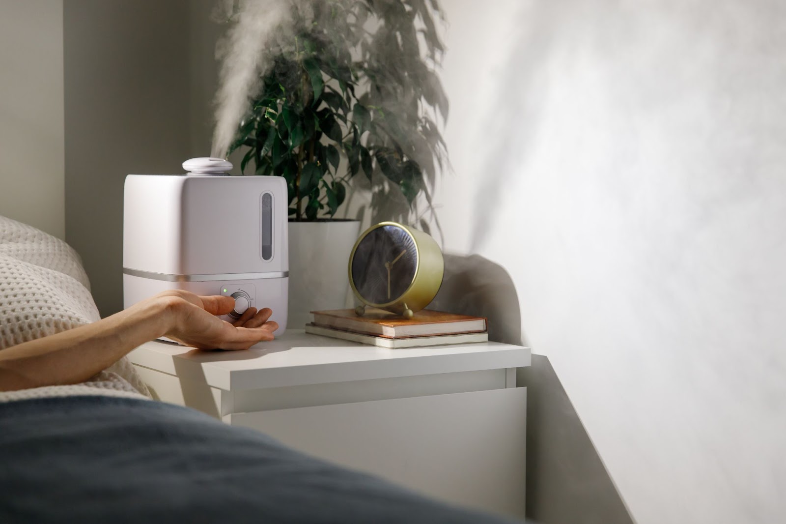 A person reaching from out of the frame to turn on a humidifier that rests on their bedside table, sitting next to an alarm clock and a plant.