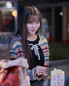 This contain an image of AKMU Suhyun' standing in front of a table with popcorn and bags on top of it