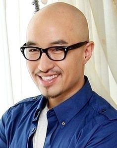 This contains an image of Hong Seok Cheon with glasses and a blue shirt smiles at the camera while standing in front of a window