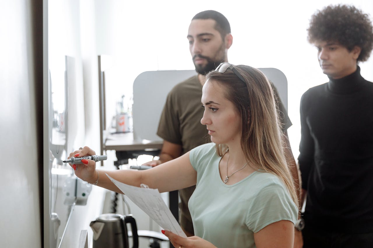 A focused woman holding papers and writing on a whiteboard in a modern office setting, with two attentive male colleagues standing behind her, discussing strategies related to board portal software.