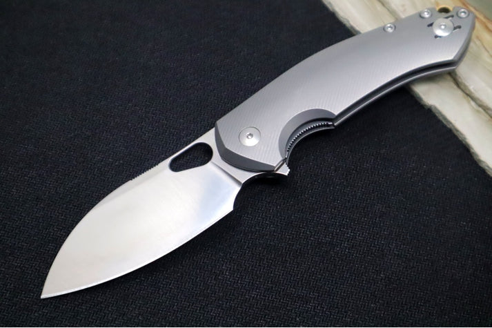 An image showing the GiantMouse ACE Biblio folding knife on a solid black and wood background. The knife features a drop-point blade and a full titanium handle.