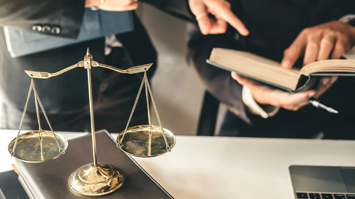 (Close-up of justice scales with two attorneys reviewing legal text in the background—symbolizing legal action and advocacy in civil rights cases.)
