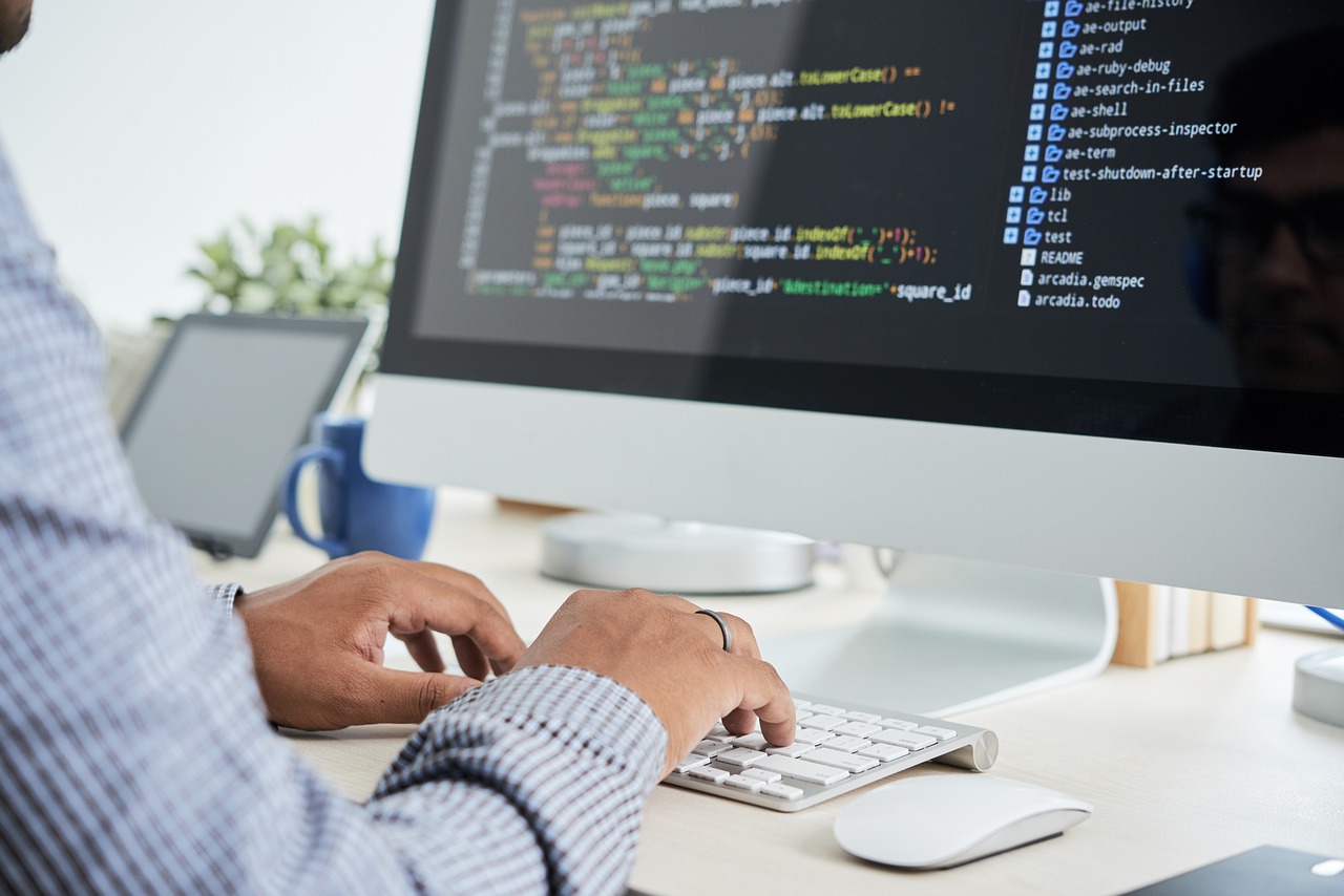 A close-up of a person's hands typing on a keyboard with lines of code displayed on a large computer screen.