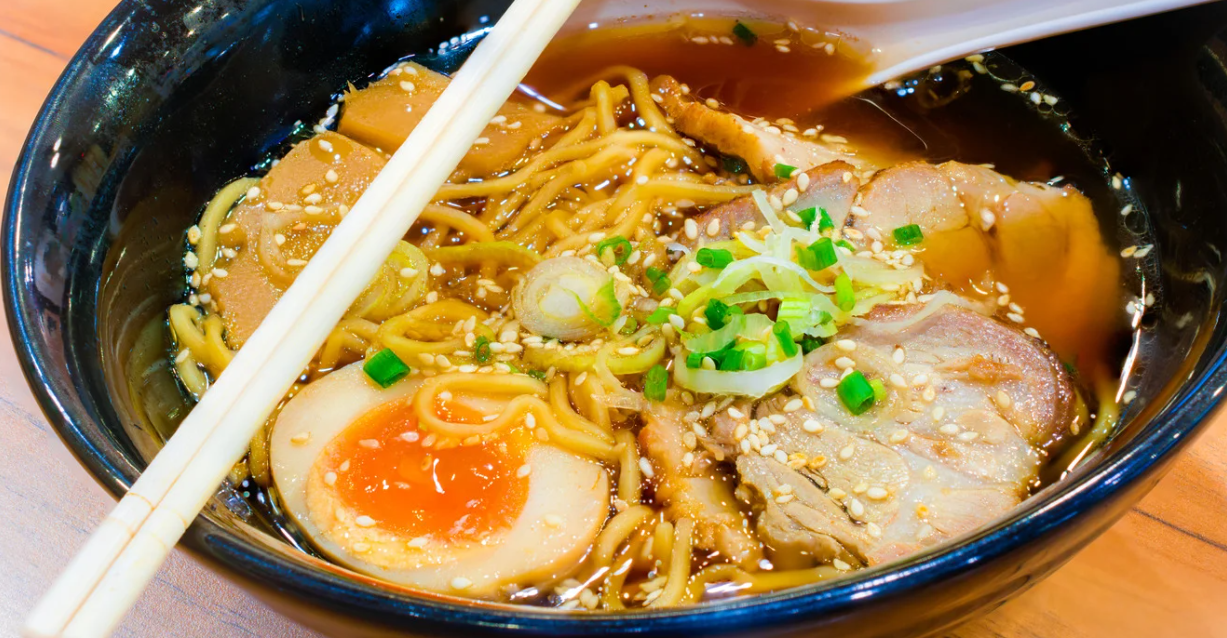 (Close-up van een kom ramen met in soja gemarineerd ei, plakjes varkensvlees, bosuitjes, bamboescheuten en sesamzaadjes in een rijke bouillon, geserveerd met eetstokjes.)