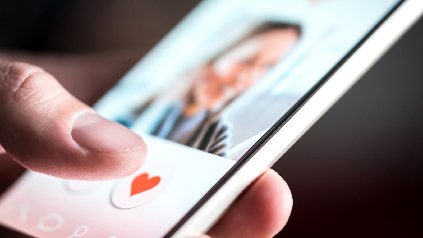 Phone screen in a hand, it is blurry but shows the profile of a woman and the finder hovering over the love heart icon