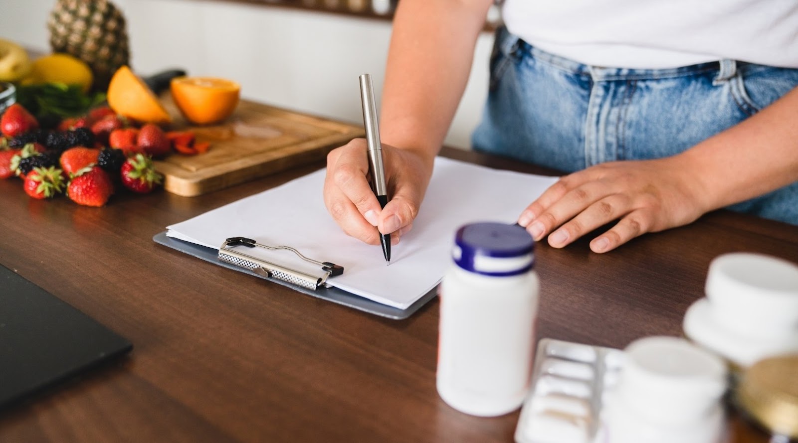 A registered dietitian writing on a clipboard near fresh fruit, supplements, and medications on a kitchen counter.