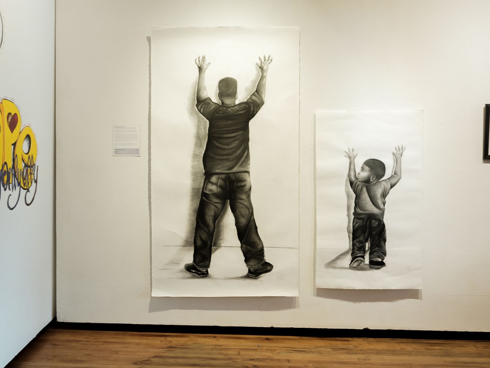 Image: Suspects by Mike Sullivan installed at exhibition More Beautiful, More Terrible: Human of Life Row.  On a gallery wall, two pieces of paper hung next to each other, the left one being about double the size of the right one. On the left paper is an illustration of a Black man standing with his hands up, facing towards the wall. He wears a black t-shirt and jeans. On the right paper is an illustration of a Black child in the same position but they're looking to the left instead of against the wall. Photo by Olivia Mikolai Ridge, 2024.