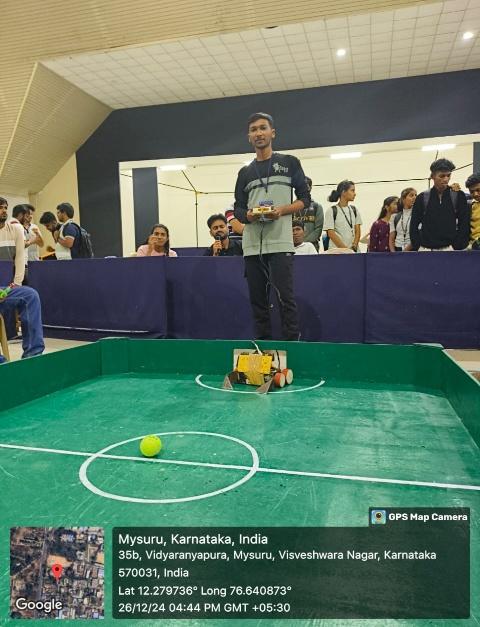 Student presenting a robot soccer model at a robotics event in Mysuru, Karnataka, with a mini football field setup and spectators watching in the background, captured with GPS map camera on December 26, 2024.