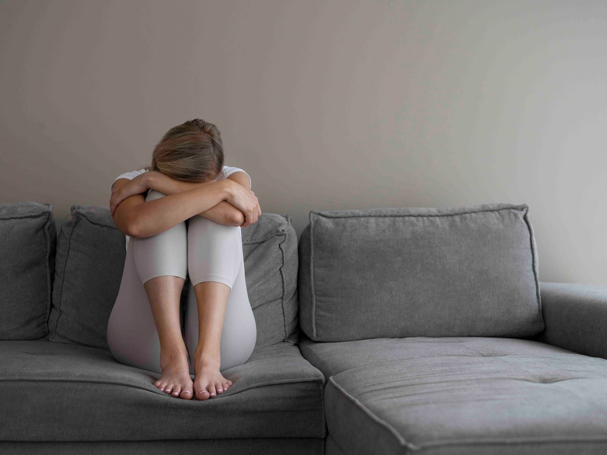 Depression, sadness, and emotional distress are symbolised by a woman sitting alone on a grey couch with her head resting on her arms and her knees pulled to her chest.