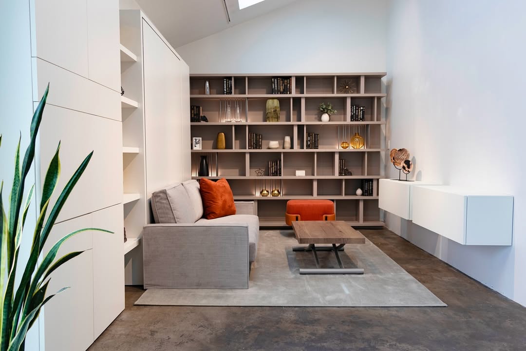 Modern multifunctional library with built-in Murphy bed, light gray sofa, open shelving and floating storage units.