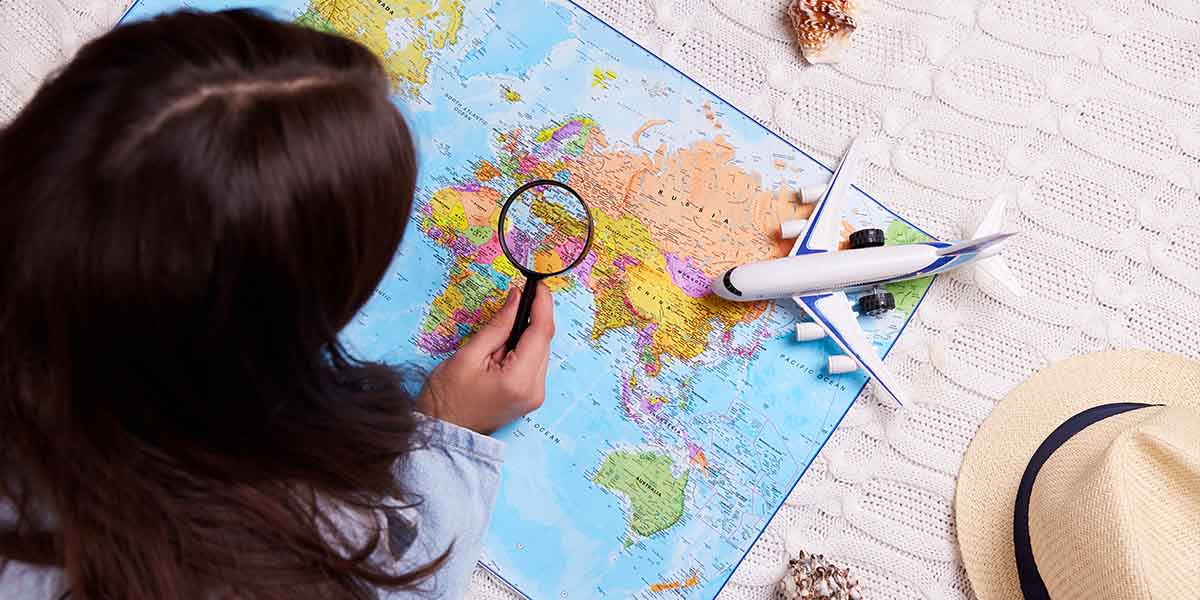 Person looking at a world map with a magnifying glass; a toy airplane, hat, and seashells are beside the map