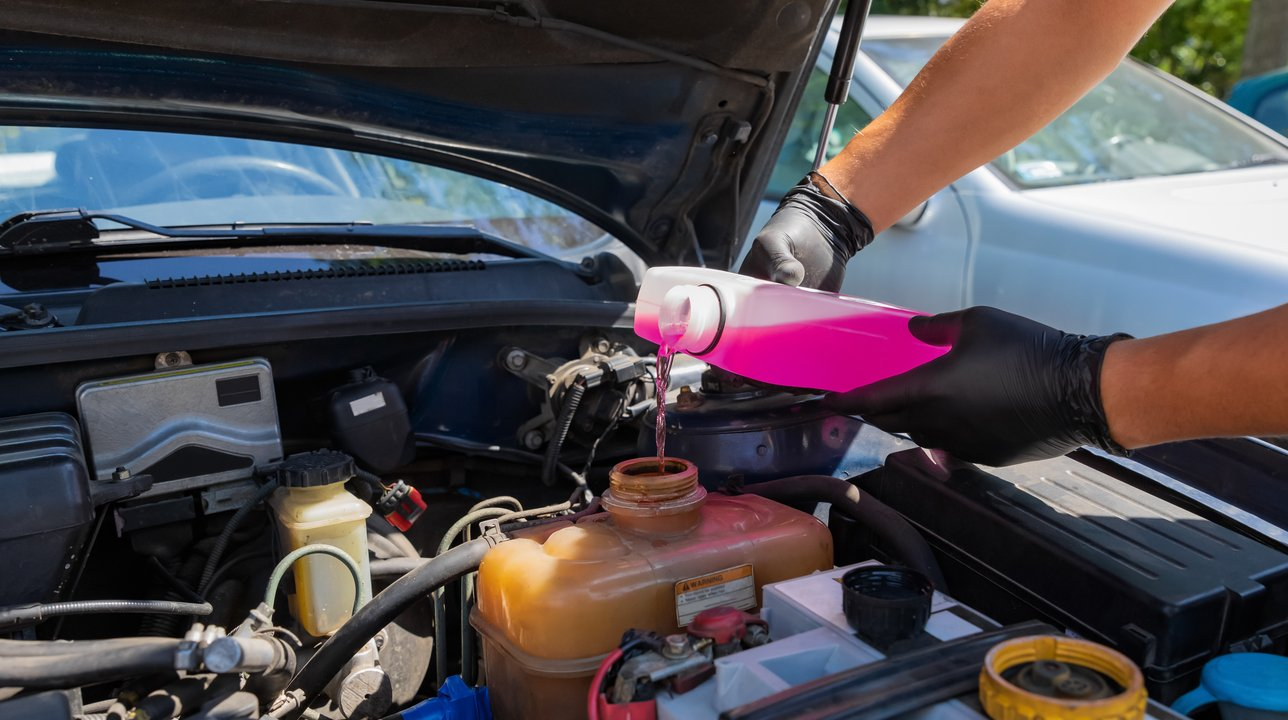 Dois braços masculinos vestindo luvas pretas segurando um recipiente com líquido rosa despejando dentro de um reservatório na parte interna do carro.