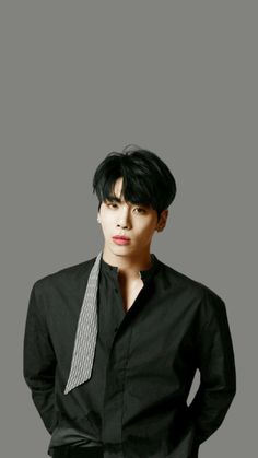 This contains an image of JONGHYUN  wearing a black shirt and tie standing in front of a gray background