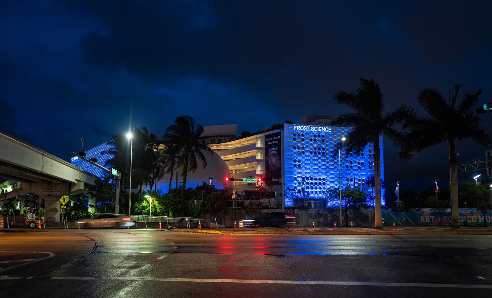 Nighttime view of the Frost Museum of Science in Miami, with its futuristic blue-lit facade, palm trees and surrounding city streets under a dark sky.
