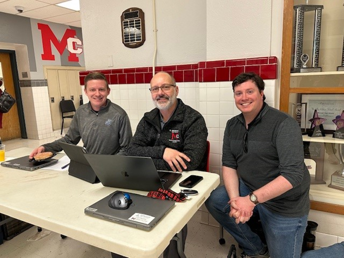 Three men seated at a table with computers. 
