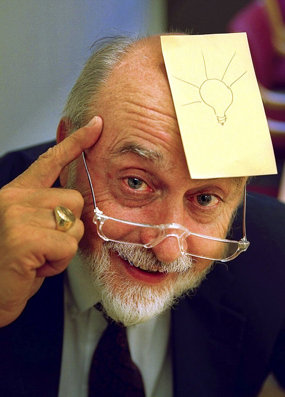 An older gentleman with glasses and a beard has a large yellow post-it note on this balding head. The post-it note has a light bulb drawn on it.